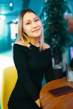 Beautiful female is sitting at table in lounge bar with mobile phone. Pretty woman in black dress in restaurant posing at camera. Party and leisure time at cafe. Portrait of smiling girl.