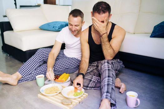 Gay couple eating together sitting on their living room floor. Homosexual lifestyle concept.