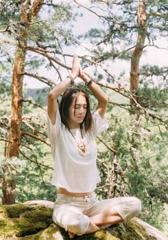 Stylish beautiful young woman practicing yoga on cliff in summer forest outdoor.