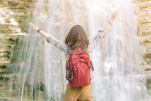 Happy backpacker explorer young woman standing with raised arms in front of waterfall on sunny summer day outdoor, rear view.