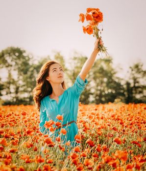 Smiling beautiful young woman holding bouquet of red poppies over her head in flower meadow in summer outdoor.