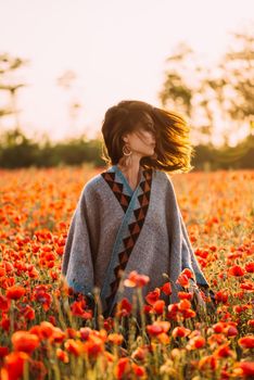 Boho stylish beautiful brunette young woman walking in red poppies meadow on sunny summer day.