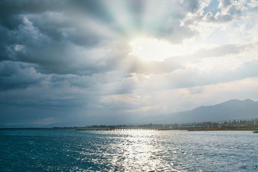 Sunny sky with clouds at sea. Resort town and mountains on horizon. Morning sun over blue ocean. Beautiful water highlights. Travel, summer vacation, tourism.
