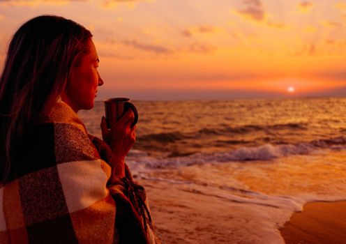 Smiling young woman wrapped in plaid relaxing with cup of beverage on beach near the sea at sunset.