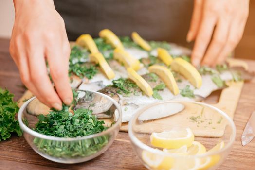 Female hands preparing fish trout with lemons and greenery on wooden table in the kitchen.