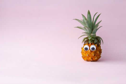 Fresh yellow funny eco pineapple with green leaves and with googly eyes on pink background on the right, no people, enough place for text. .