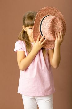 Girl in stylish summer clothes. A girl with ponytails in a pink blouse, white trousers covers her face with a straw hat. Portrait of a serious young child