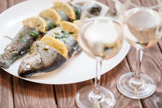 Simple fish dish with lemons and greenery on a white plate on wooden table with two glasses of white wine.