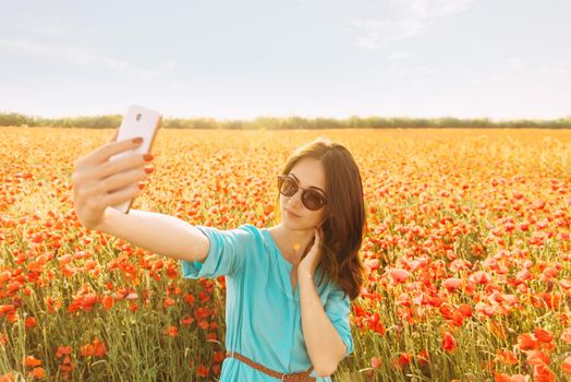 Beautiful young woman taking selfie with smartphone on background of red poppies meadow on sunny summer day.
