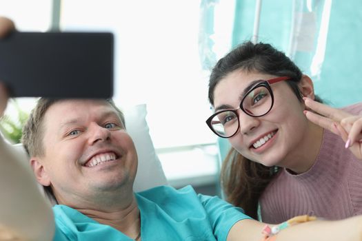 Portrait of smiling young woman and hospital patient taking selfie on mobile phone. Cheerful family picture. Modern clinic room for sick people. Medicine and recovery concept