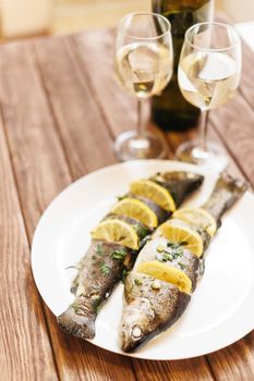 Fish dish with lemons and greenery on a plate on wooden table and glasses of white wine for couple.