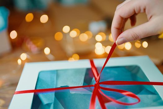 Close-up of girl's hand pulling red ribbon from gift. Bokeh lights in background. Unpacking box on wooden table. Festive concept of Christmas or Birthday.