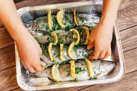 Unrecognizable woman preparing fish trout with lemons and greenery on foil tray, view of hands.