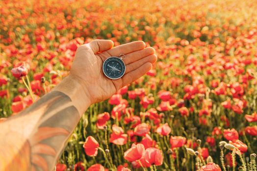Man’s hand with travel compass on background of red poppies meadow, point of view.