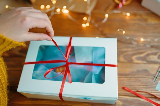 First-person view of hand in knitted sweater opening Christmas surprise. Woman pulls ribbon on gift box. Wooden table with garland. New Year's holiday mood.