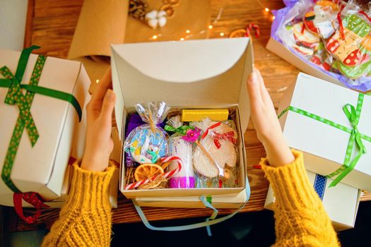 Women's hands in knitted sweater open Christmas gift. Gingerbread with icing, candle with ornamental, souvenir made of polymer clay and decorations in box. Boxes on wooden table.