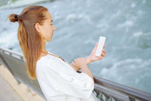 Beautiful young woman standing with smartphone on embankment near the river.