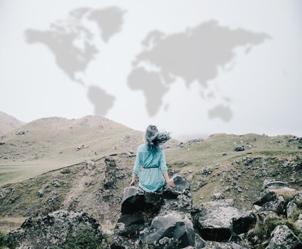 Young woman sitting on rocky mountain outdoor, rear view. Map of world is painted on sky. Concept of travel.