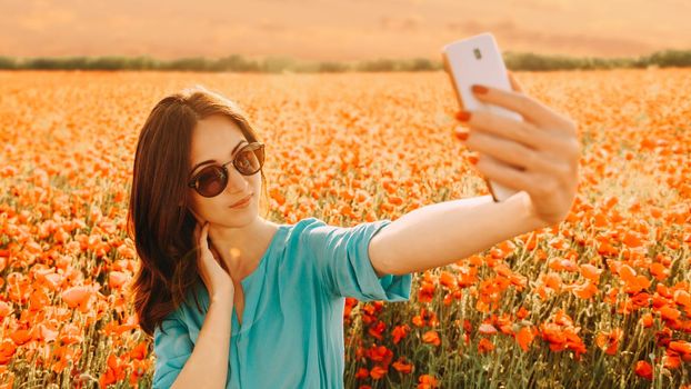 Beautiful girl making selfie with smartphone on background of poppies flower meadow in summer at sunset.