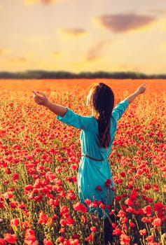 Happy young woman standing with raised arms in red poppies meadow at sunset.