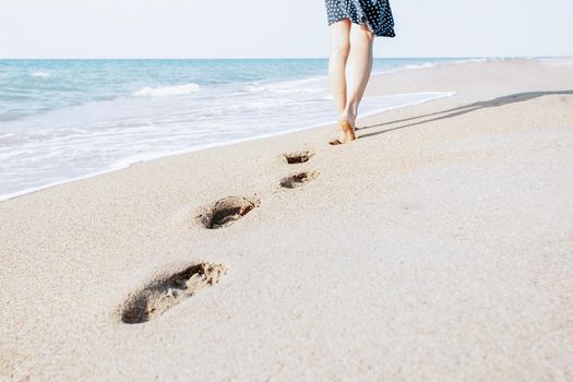Unrecognizable barefoot woman walking on beach leaving footprints in the sand near sea, view of legs.