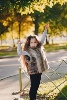 Stylish baby girl 4-5 year old wearing boots, fur coat standing in park. Looking at camera. Autumn fall season.