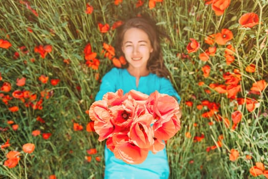 Happy young woman lying on flower meadow and giving a bouquet of red poppies, top view. Focus on flowers.