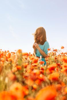 Brunette young woman standing in red poppies meadow and sniffing a flower on sunny summer day.