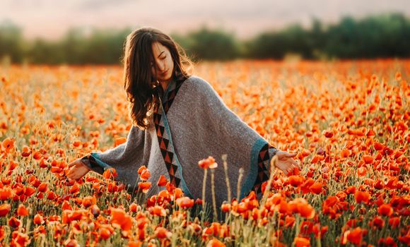Beautiful boho style young woman walking in flower meadow and touching red poppies in summer outdoor.