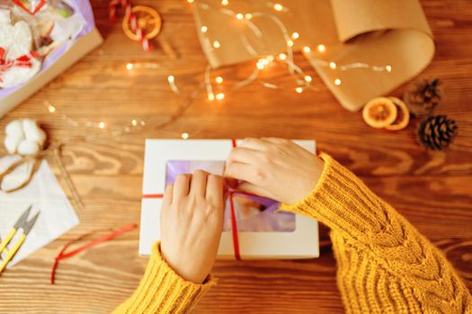 Top view of girl's hands in knitted orange sweater tie red ribbon bow on white gift box. Garland lights, scissors, pine cone and decorations in background. New Year's mood. Surprise for Christmas.