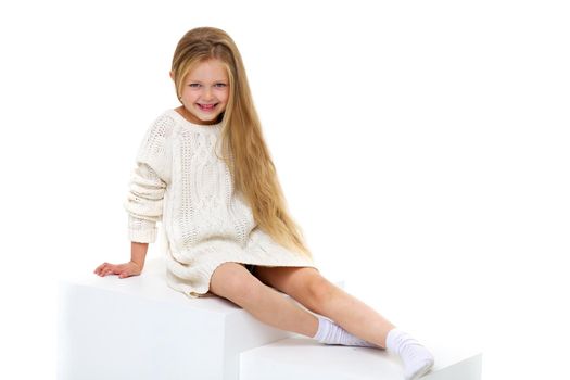 Cute little girl wearing warm knitted dress sitting on white staircase. Happy smiling long haired girl posing in studio against white background. Happy childhood concept