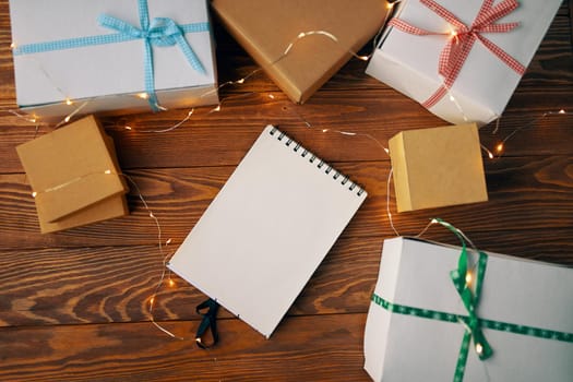 Wooden table with gift boxes blank spiral bound notepad and garland. Copy space for writing wishes. Concept of Christmas new year or birthday. Boxes are tied with colored ribbons.