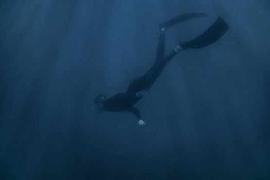 Young man freediver wearing in wetsuit and flippers swimming underwater in blue deep sea.