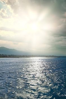 Seascape at dawn. Glare on the surface of ocean. Pier, trees and mountains in background. Resort town. Travel concept.