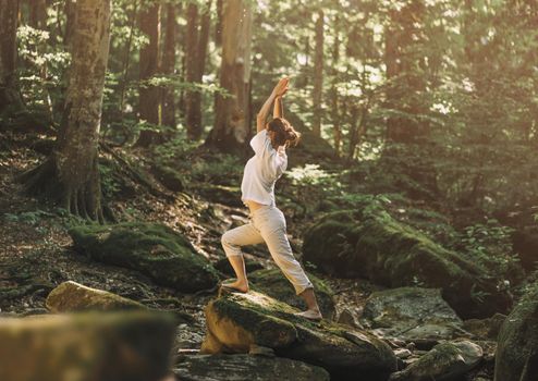 Young woman wearing in white clothing practicing yoga in pose of warrior in summer forest outdoor.