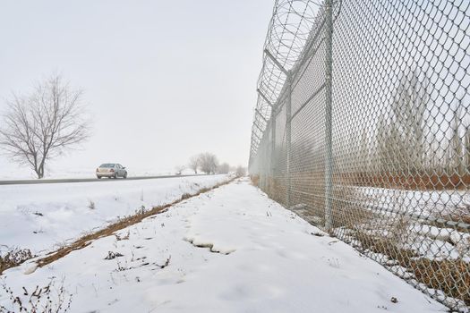 Barbed wire fence on border in winter. Private secured object near highway. Maximum security detention facility. Car goes on road. Closing for quarantine. Unauthorized entry is prohibited.