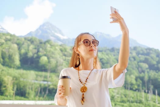 Smiling young woman with cup of coffee taking selfie with smartphone on balcony on background of summer mountains.