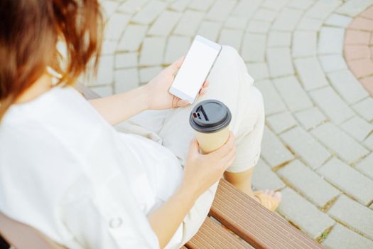 Unrecognizable young woman sitting with cup of coffee and smartphone on wooden bench.