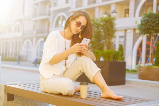 Stylish beautiful young woman sitting on wooden bench with smartphone and cup of coffee in street outdoor.