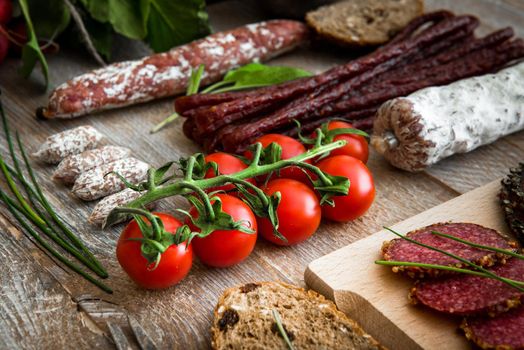 A bunch of cherry tomatoes with salami sausages, bread slices, greens on a wooden textured background