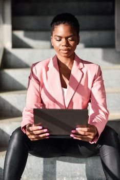 Black businesswoman sitting on urban steps working with a laptop computer. African american female wearing suit with pink jacket.