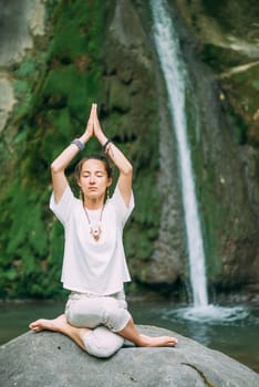 Stylish young woman practicing yoga on stone near the waterfall in summer.