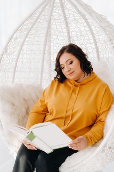 a woman in an orange hoodie sits in an unusual chair with a book in her hands.