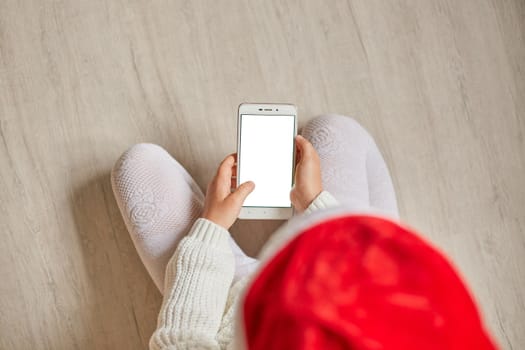 Top view of little child with smartphone with blank screen in hands hands, kid wearing santa hat, white pullover and tights, Copy space for advertisement.