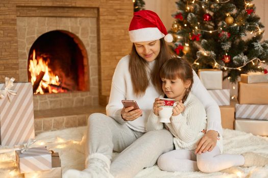Attractive female wearing white jumper and santa hat sitting with her daughter on floor, looking at smart phone screen in mom's hand, posing in living room with New Year decoration.