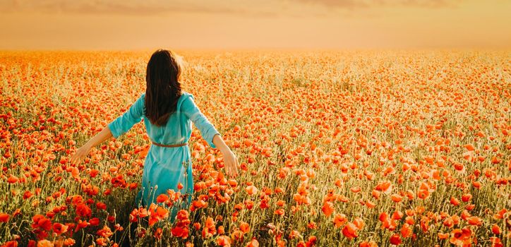 Brunette young woman wearing in blue dress walking in poppies flower meadow at sunset.