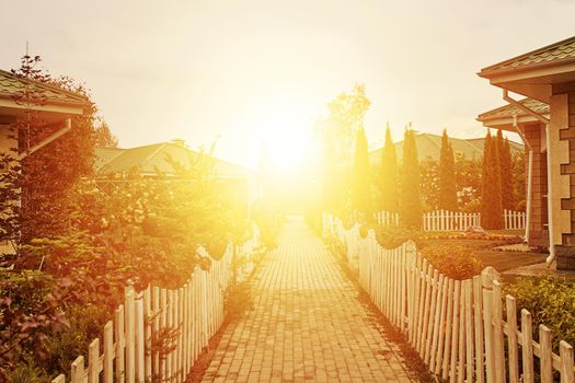 Sidewalk between cottages and gardens to rising sun. Small fences around road. Nice houses and trees. Summer landscape. Bright sunlight at end of street.