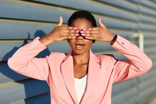 Funny black woman, model of fashion, standing on urban wall covering her eyes with her hands. African american female wearing suit with pink jacket with sunset light.