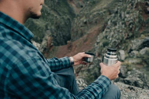 Explorer young man sitting with thermos in summer mountains outdoor, close-up.