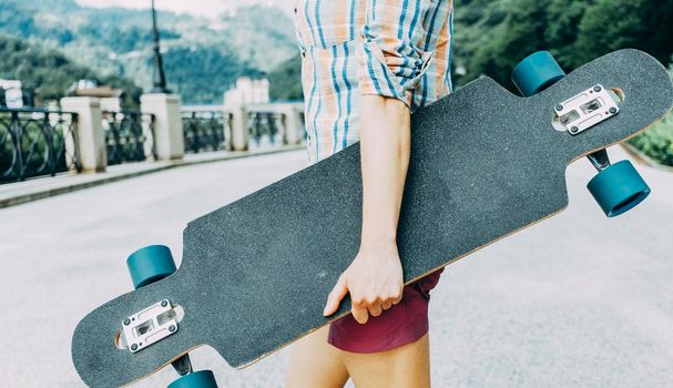 Unrecognizable teenager standing with longboard on street. Copy-space on surface of board.